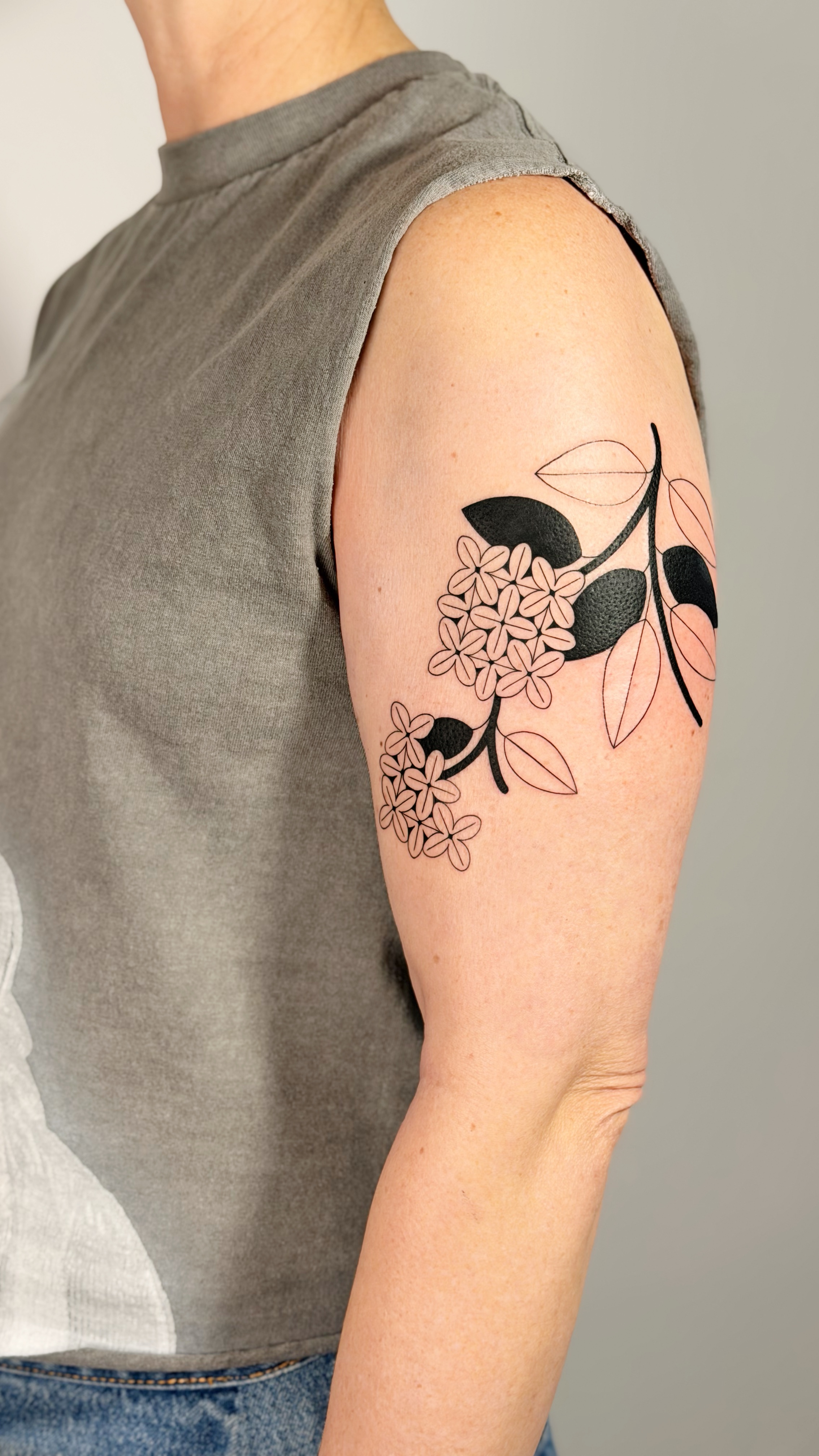 Blackwork shoulder tattoo of a botanical flower branch with bold leaves and blossoms — fine line work and graphic floral illustration by Jane Again Tattoo, Mexico City.