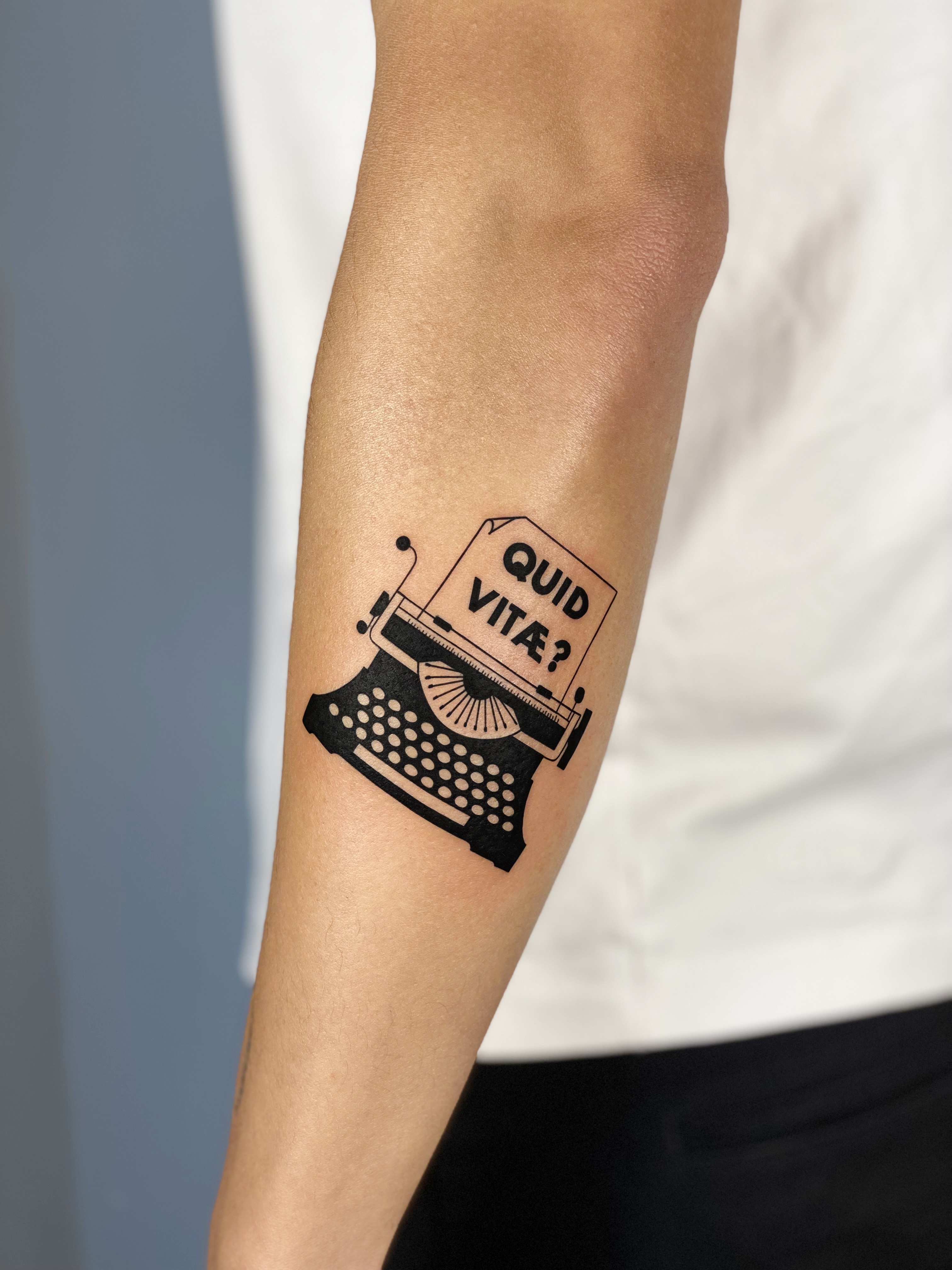 Blackwork forearm tattoo on dark skin of a vintage typewriter with the words 'Quid Vitae?' — bold graphic line work and illustrative lettering by Jane Again Tattoo, Mexico City.