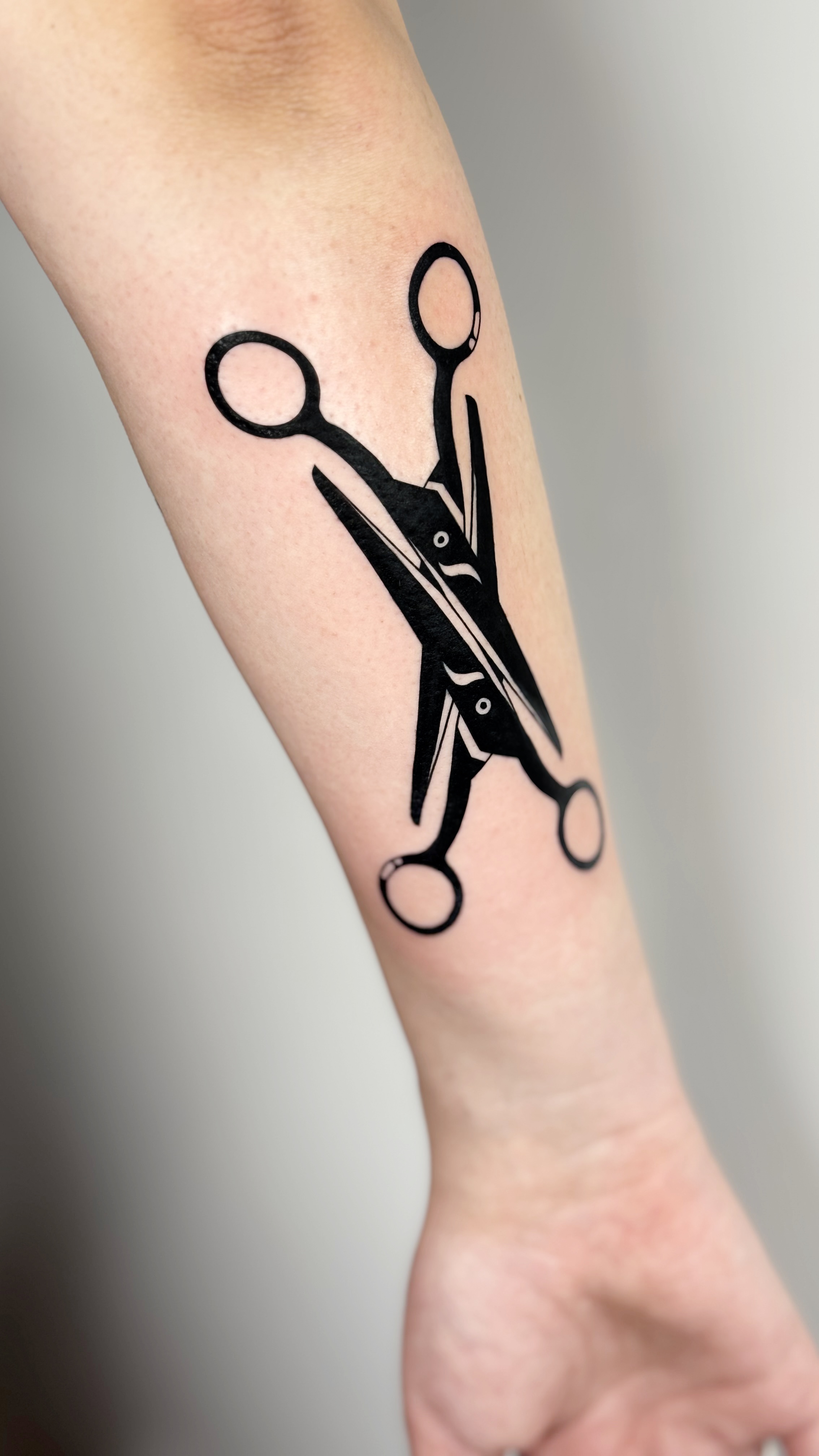 Blackwork queer tattoo of two intertwined scissors on the forearm — lesbian iconography in bold graphic minimalist line work and fine line illustration by Jane Again Tattoo, Mexico City.