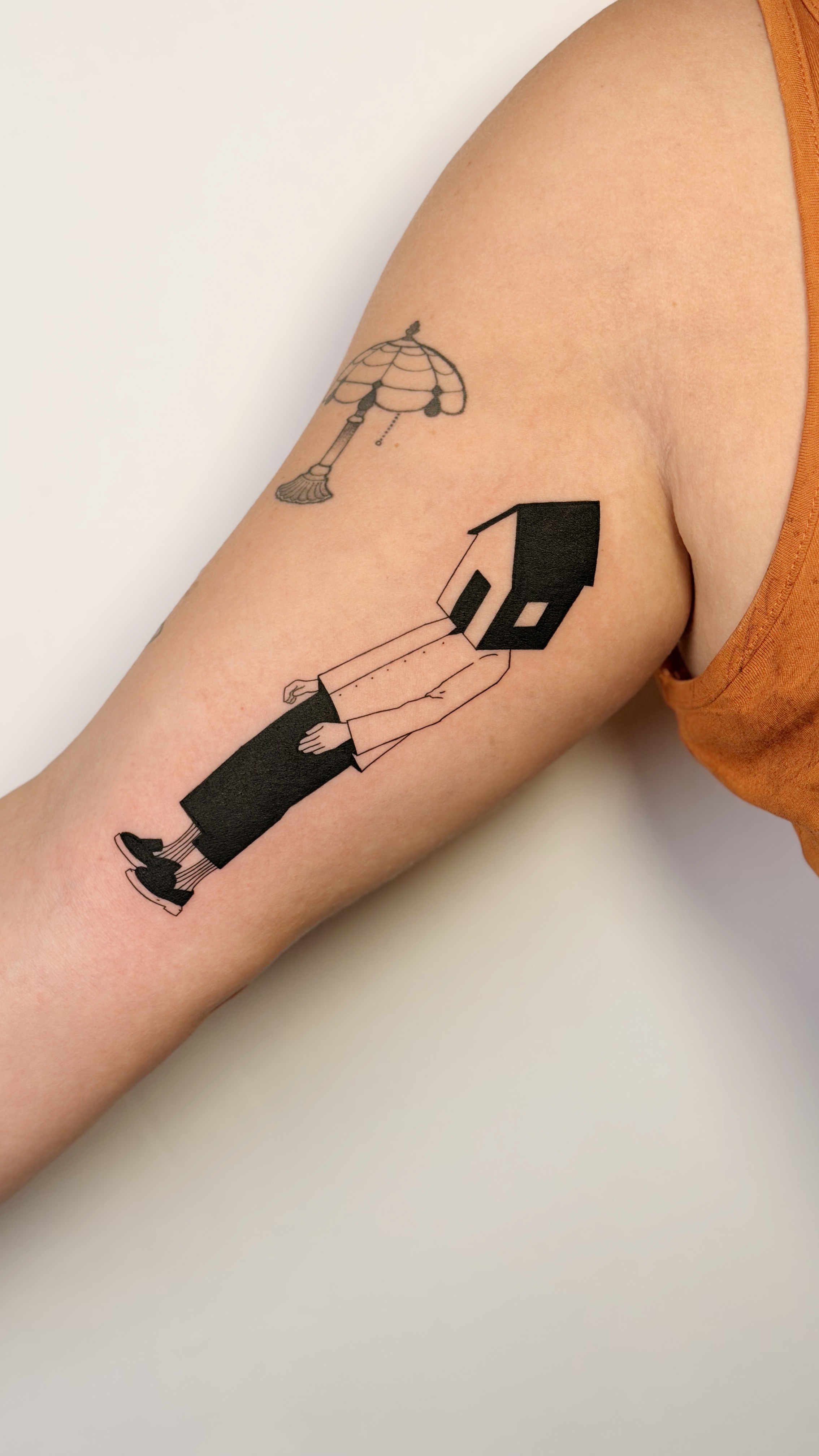 Blackwork arm tattoo of a figure with a house-shaped head beside a Tiffany-style lamp — surreal illustrative line work and graphic narrative by Jane Again Tattoo, Mexico City.