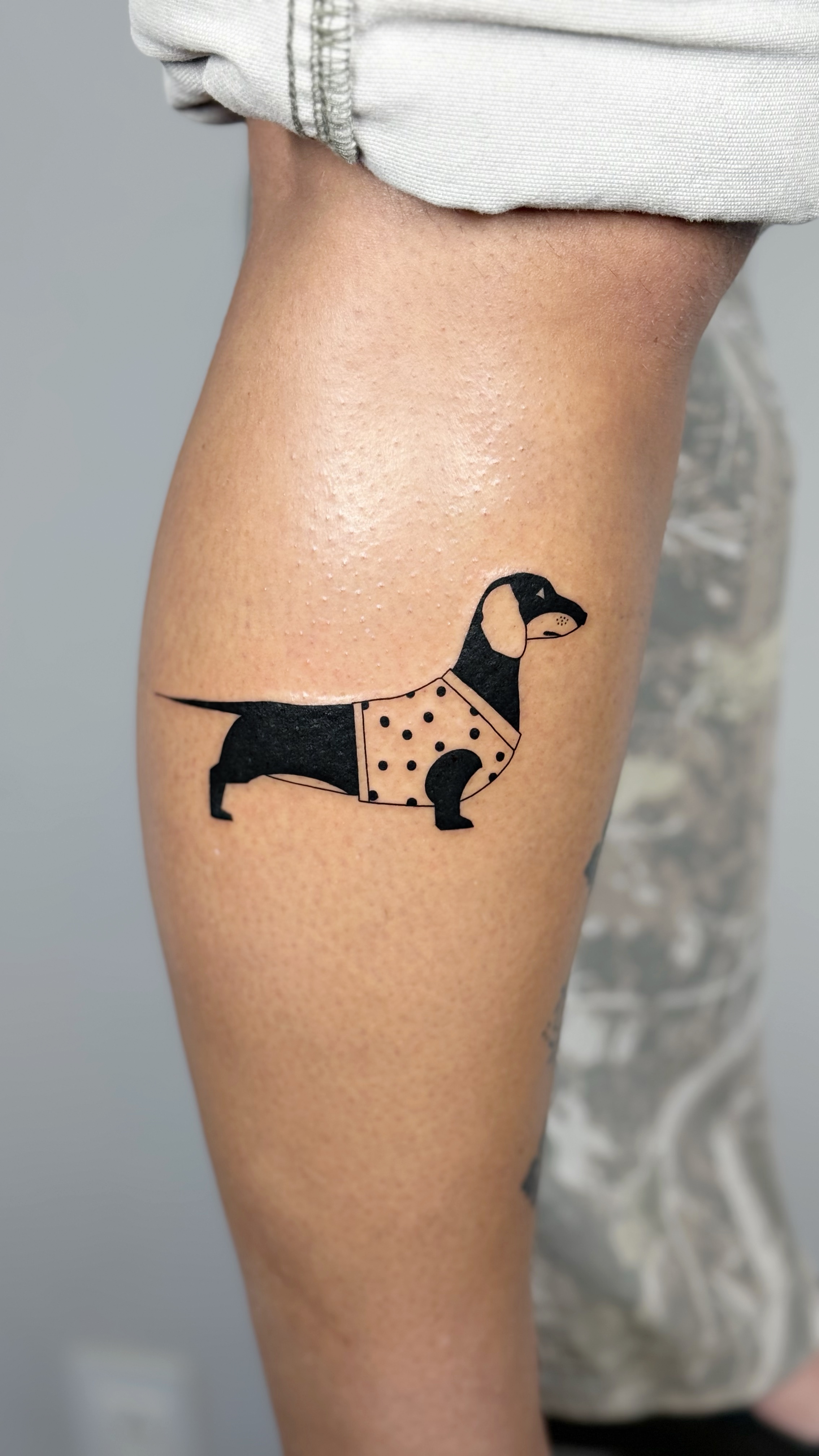 Bold blackwork dachshund tattoo styled as a Mexican lotería card on the calf — graphic illustrative line work and Mexican folk-art composition by Jane Again Tattoo, Mexico City.