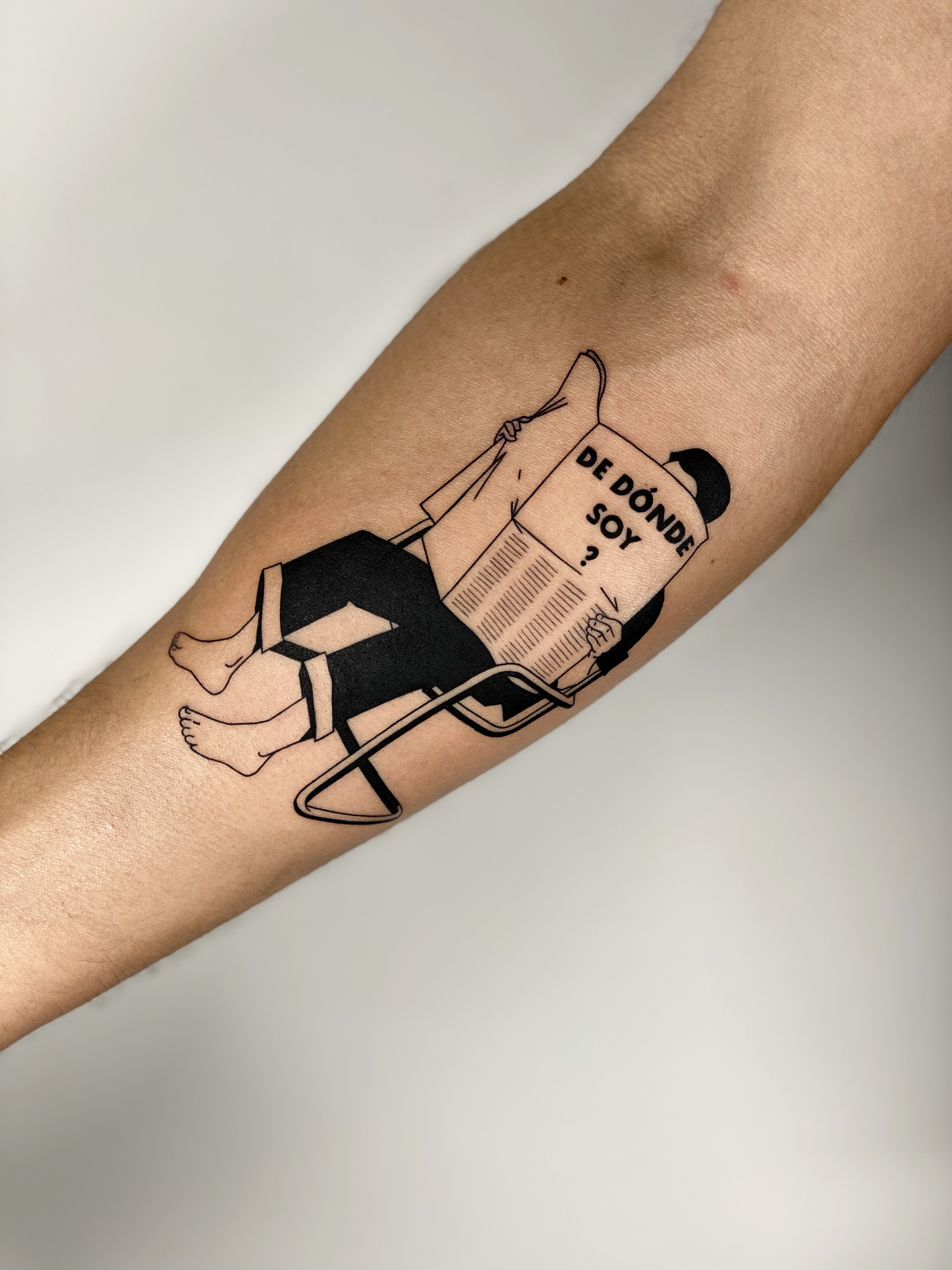 Blackwork arm tattoo on dark skin of a person reading a newspaper titled 'De Dónde Soy' — illustrative graphic line work and narrative composition by Jane Again Tattoo, Mexico City.
